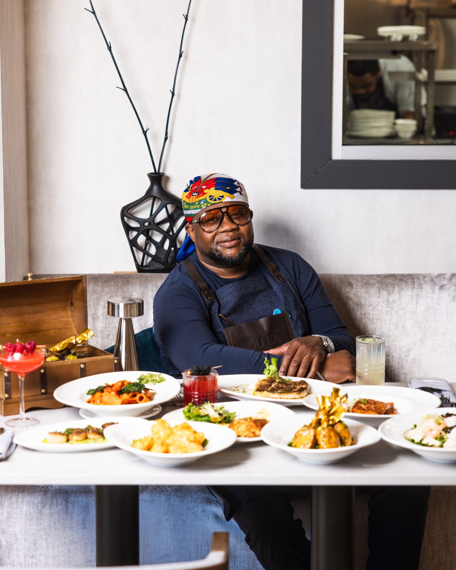 Chef Duke of OU LA restaurant, wearing a Haitian flag bandana and stylish sunglasses, sits proudly at a table adorned with an array of his signature Caribbean fusion dishes. The spread includes grilled lamb chops in a wooden treasure chest, seafood pasta, seared scallops, crab cakes, and handcrafted cocktails. The open kitchen in the background adds to the dynamic dining experience at OU LA.